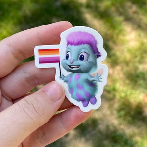 Könnte beinhalten: Ein Aufkleber mit einer blauen und violetten Comic-Fee mit pinken Haaren, die eine lesbische Pride-Flagge hält. Die Flagge ist rot, orange, gelb und pink.