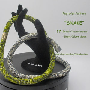 May include: A green and white peytwist beaded necklace with a single column seam. The necklace is 17 beads in circumference and is called "SNAKE". The necklace is on a black stand. Peytwist Pattern "SNAKE" 17 Beads Circumference Single Column Seam www.etsy.com/shop/ShinyBeadArt © Jane Junckert