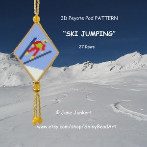May include: A beaded pendant with a ski jumping design. The pendant is diamond-shaped, featuring a skier in red, yellow, and green on a blue background. The background shows a snowy mountain scene. Text on the image reads "SKI JUMPING".