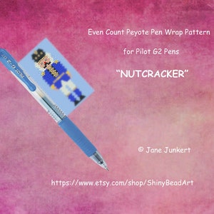 May include: A blue pen with a white label that says "PILOT G-2 07" is lying on top of a blue and white bead pattern of a nutcracker. The pattern is labelled "Even Count Peyote Pen Wrap Pattern for Pilot G2 Pens" and "NUTCRACKER". The image also includes the text "https://www.etsy.com/shop/ShinyBeadArt" and "Jane Junkert".
