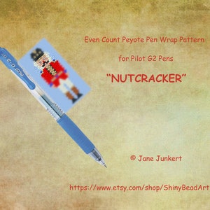 May include: A blue pen with a white label that says "PILOT G-2 07" is lying on top of a printed pattern for a beaded pen wrap. The pattern is of a nutcracker with a red and white costume. The text "Even Count Peyote Pen Wrap Pattern for Pilot G2 Pens "Nutcracker" Jane Junkert https://www.etsy.com/shop/ShinyBeadArt" is printed below the pattern.
