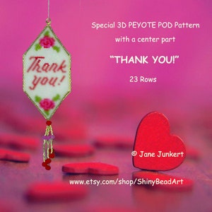 May include: A beaded ornament with the text "Thank you!" in red letters, framed by pink roses and green leaves. The ornament hangs from a gold-colored loop and has a beaded tassel. A red heart is in the foreground.