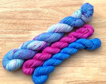 Könnte beinhalten: Drei Knäuel Garn in verschiedenen Farben: ein hellblaues mit weißen Sprenkeln, ein leuchtendes Pink und ein solides Dunkelblau.