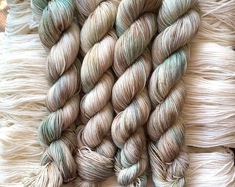 May include: Four skeins of yarn in shades of beige, brown, and green. The yarn is arranged in a criss-cross pattern.