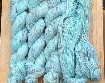 May include: Five skeins of light blue yarn with flecks of color. The yarn is arranged in a pile on a wooden surface.