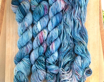 May include: Four skeins of blue, green, and white yarn with speckles of color. The yarn is arranged in a criss-cross pattern.