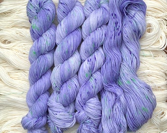 May include: Four skeins of lavender yarn with flecks of green and purple. The yarn is arranged in a fan shape.