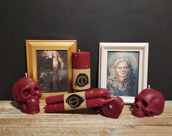 May include: Three red skull-shaped candles and three red pillar candles with gold accents. The candles are arranged on a wooden surface in front of two framed pictures. One picture features a woman in a flowing gown and the other picture features a woman with a half-skull face.