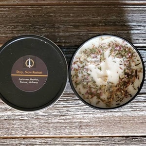 May include: A black candle tin with a label that reads "Stop, Now Restart" and lists the ingredients: Agrimony, Heather, Yarrow, Mulberry. The candle is white and has dried herbs and flowers on top.