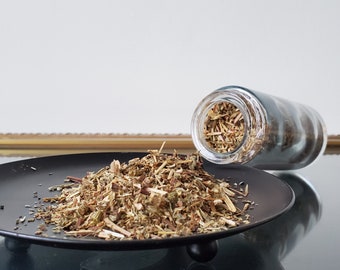 May include: A black plate holds a pile of dried herbs, with a clear glass jar tipped over beside it. The herbs are a mix of green, brown, and tan, with visible stems and leaves. The jar contains similar herbs.