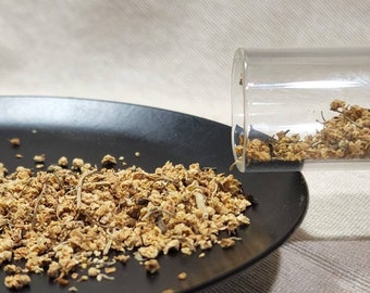 May include: A glass tube filled with dried flower buds is being poured onto a black plate. The flower buds are a light brown color and have a slightly fuzzy texture.