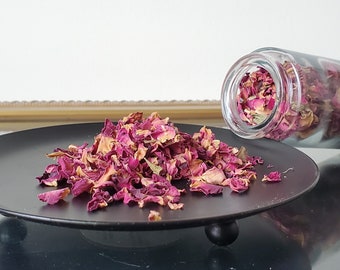 Puede incluir: Un plato negro con un montón de pétalos de rosa secos. Los pétalos son una mezcla de rosa y rojo. Un frasco de vidrio con más pétalos de rosa está volcado sobre el plato.