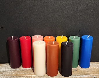 May include: Ten pillar candles in various colors, including purple, red, cream, brown, black, green, and blue. The candles are arranged in a row on a wooden surface.