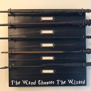 May include: A wooden wall-mounted wand holder with six wands displayed. The holder has a black finish and features a white inscription that reads "The Wand Chooses The Wizard".