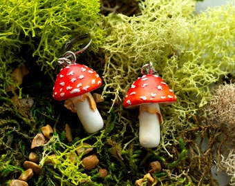 Op de afbeelding: Een paar rode en witte paddenstoel oorbellen met zilveren haakjes. De paddenstoelen zijn gemaakt van polymer clay en hebben een realistische uitstraling. De oorbellen zijn geplaatst tegen een achtergrond van groen mos.
