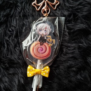 May include: A white character with black hair and a red mark on their forehead sits on a pink and orange swirl lollipop. The lollipop is attached to a white stick with a yellow bow with white polka dots. The lollipop is packaged in clear plastic with a gold bow and chain.