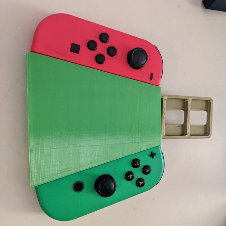 May include: A green and pink Nintendo Switch console with a game cartridge slot on the side. The console is in a stand that allows it to be used in tabletop mode.