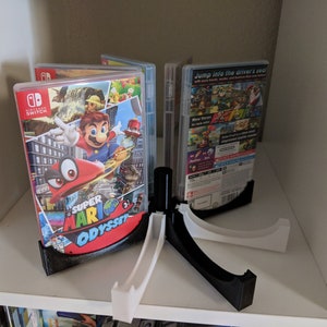 May include: A black and white plastic stand holds multiple Nintendo Switch game cases. The stand is designed to hold the cases upright. The game cases are for Super Mario Odyssey and Mario Kart 8 Deluxe.