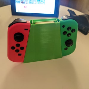 May include: A green and pink Nintendo Switch controller with black buttons. The controller is in a stand that is made of green plastic.