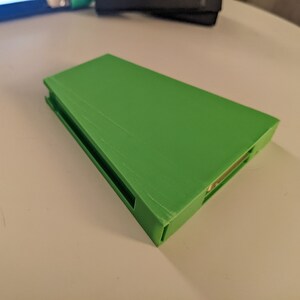 May include: A bright green 3D printed case with a slot for a small device. The case has a smooth, slightly textured surface.