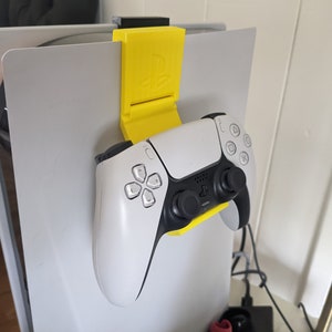 May include: A yellow 3D printed controller holder attached to a white console. The holder is designed to hold a white PlayStation 5 DualSense controller.