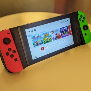 May include: A Nintendo Switch console with a red and green controller. The screen shows the game "Paper Mario: The Origami King" and other game icons.