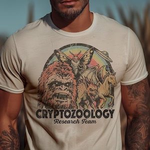 Op de afbeelding: Crèmekleurig T-shirt met een kleurrijke afbeelding van cryptiden, waaronder een Bigfoot, een mot en een gevleugeld wezen, met de tekst "CRYPTOZOOLOGY Research Team". Het shirt wordt gedragen door een persoon met tatoeages.
