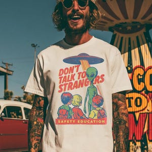 Könnte beinhalten: Weißes T-Shirt mit einem Retro-Grafikdesign einer Alien-Entführungsszene und dem Text "DON'T TALK TO STRANGERS SAFETY EDUCATION". Die Person trägt einen Aluhut und eine Sonnenbrille.