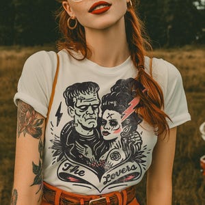 Pu&ograve; includere: T-shirt bianca con grafica in bianco e nero di una coppia e la scritta "The Lovers". La persona indossa occhiali con montatura trasparente, orecchini a cerchio dorati e ha i capelli rossi. Indossa anche una cintura arancione e ha un tatuaggio sul braccio.
