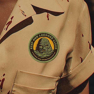May include: A woman wearing a tan short-sleeved shirt with blood-like stains and a patch that reads "Keep Our Lagoons Clear." The shirt has a buttoned front and a pocket. A creature is visible in the background.