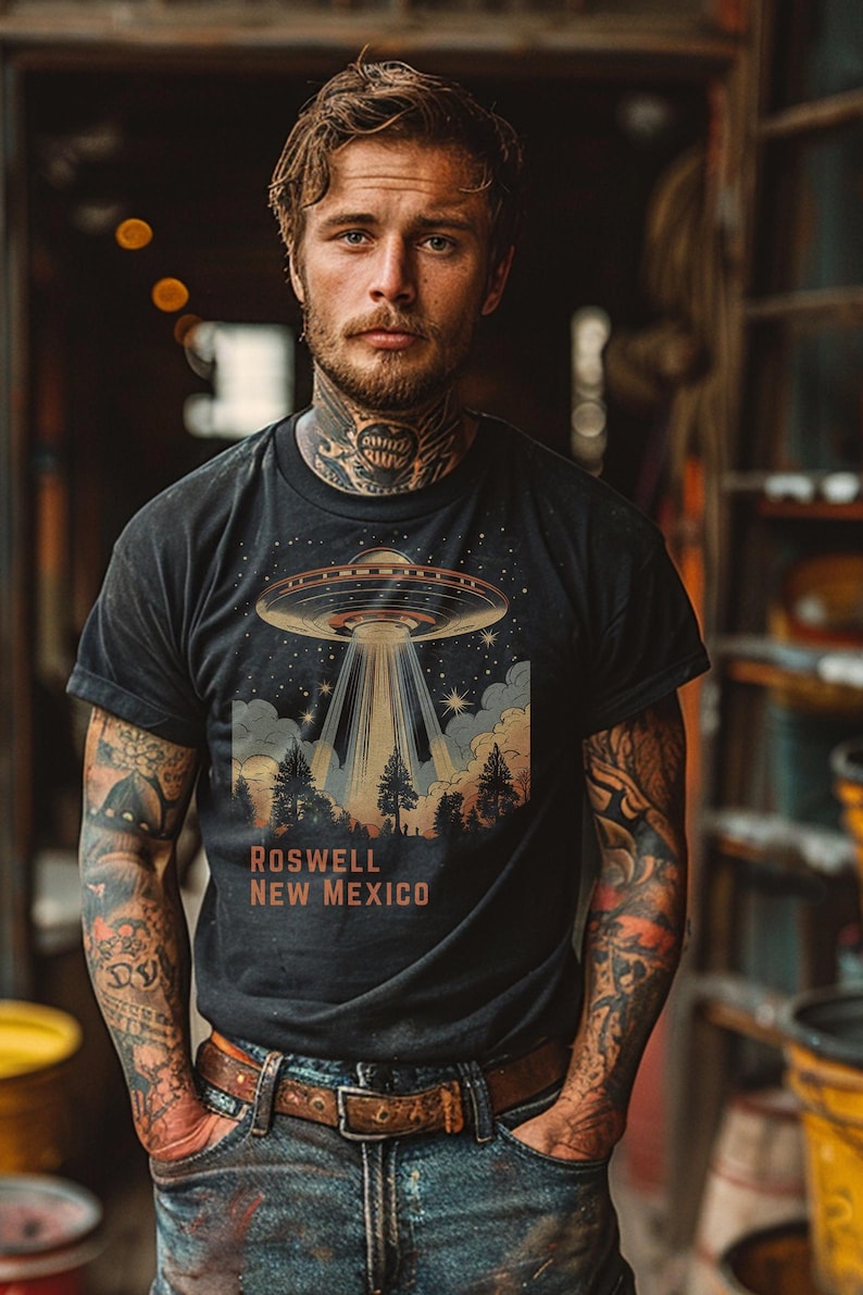 Puede incluir: Camiseta negra con un dise&ntilde;o retro de un OVNI que proyecta un rayo de luz sobre un bosque, con el texto "ROSWELL NEW MEXICO" en naranja. La camiseta se lleva con vaqueros azules y un cintur&oacute;n marr&oacute;n.