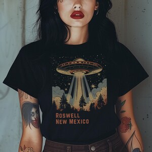 Puede incluir: Camiseta negra con una ilustraci&oacute;n retro de un OVNI que proyecta luz sobre un bosque, con el texto "ROSWELL NEW MEXICO" en naranja. La camiseta tiene cuello redondo.