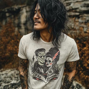 Pu&ograve; includere: T-shirt grigio chiaro con una grafica di Frankenstein e la sua sposa, un fulmine rosso e la scritta "The Lovers". La maglietta &egrave; indossata da una persona con lunghi capelli neri e tatuaggi.