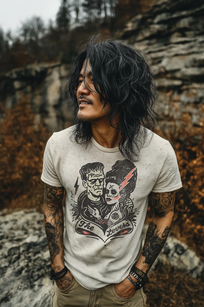 Pu&ograve; includere: T-shirt grigio chiaro con una grafica di Frankenstein e la sua sposa, un fulmine rosso e la scritta "The Lovers". La maglietta &egrave; indossata da una persona con lunghi capelli neri e tatuaggi.