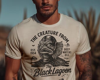 May include: Cream-colored t-shirt featuring a black and white graphic of the Creature from the Black Lagoon. The design includes the text "The Creature From", "The Black Lagoon", and "Amazon Basin". The shirt also has the text "EST. 1954" and "Deep Waters Div."