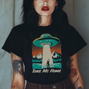 May include: Black t-shirt featuring a retro-style graphic of a UFO beaming a person up. The design includes mountains, water, and the text "TAKE ME HOME". The shirt is worn by a person.
