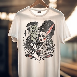 Pu&ograve; includere: T-shirt bianca con un'illustrazione in bianco e nero di Frankenstein e la sua sposa, con un fulmine rosso e le parole "The Lovers" su uno striscione. La maglietta &egrave; appesa a una gruccia di legno.