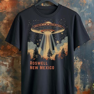 Puede incluir: Camiseta negra con un dise&ntilde;o retro de un OVNI que proyecta un rayo de luz sobre un bosque. El texto "ROSWELL NEW MEXICO" est&aacute; impreso en naranja debajo. La camiseta est&aacute; colgada en una percha negra.