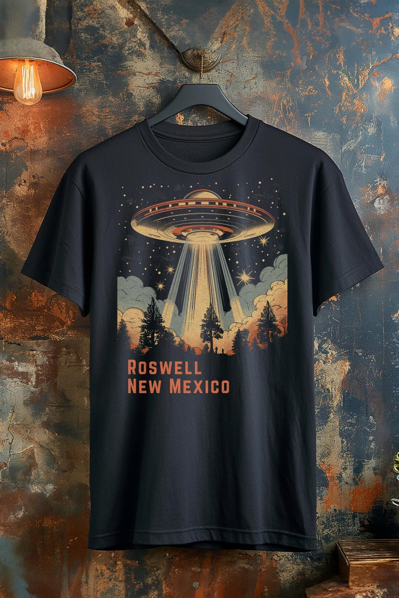 Puede incluir: Camiseta negra con un dise&ntilde;o retro de un OVNI que proyecta un rayo de luz sobre un bosque. El texto "ROSWELL NEW MEXICO" est&aacute; impreso en naranja debajo. La camiseta est&aacute; colgada en una percha negra.