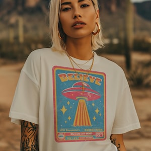 May include: White t-shirt with a retro graphic of a UFO emitting a beam of light. The graphic includes the word "BELIEVE" and the phrase "UFO Encounters". The shirt is worn by a person with blonde hair and gold jewelry.