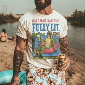 Op de afbeelding: Een crèmekleurig T-shirt met ronde hals en een afbeelding van een groen wezen in een roze zwemband met een drankje. De tekst luidt HALF MAN. HALF FISH. FULLY LIT. BLACK LAGOON FLOAT CREW. Het shirt wordt gecombineerd met een lichtblauwe zwembroek.