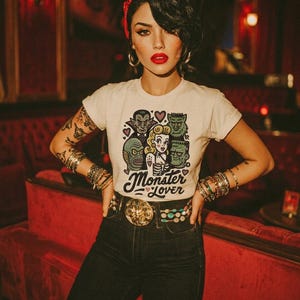 Pu&ograve; includere: Una maglietta bianca con una grafica "Monster Lover" con mostri dei cartoni animati e una donna. La persona indossa una fascia rossa, orecchini a cerchio e diversi braccialetti. Indossa anche una cintura decorativa e jeans scuri.