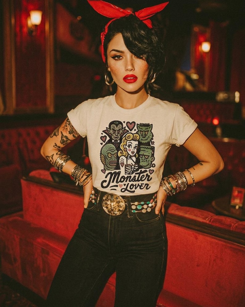 Pu&ograve; includere: Una maglietta bianca con una grafica "Monster Lover" con mostri dei cartoni animati e una donna. La persona indossa una fascia rossa, orecchini a cerchio e diversi braccialetti. Indossa anche una cintura decorativa e jeans scuri.