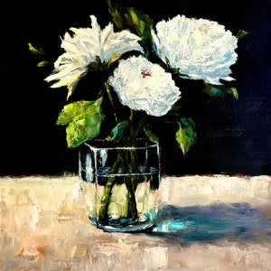 May include: White flowers in a clear glass vase on a light brown surface. The flowers are arranged in a bouquet and have green stems. The background is black.