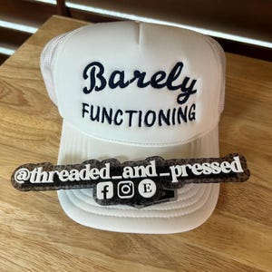 May include: White trucker hat with the words "Barely FUNCTIONING" embroidered in navy blue. A clear plastic tag with the text "@threaded_and_pressed" and social media icons rests on the hat. The hat is on a wooden surface.