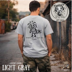 Könnte beinhalten: Hellgraues T-Shirt mit einem Cartoon-Bienen-Design und dem Text "Seemed Like a Good Idea Earlier". Das Shirt hat auch ein "Bad Ant Energy"-Logo auf der linken Brust. Das Shirt ist eine Komfortfarbe.