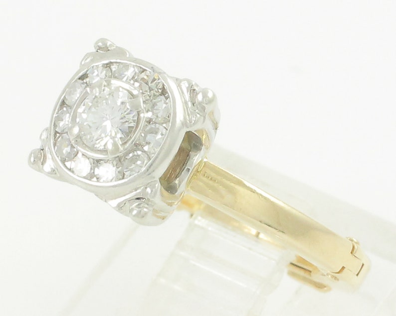 May include: A gold and white diamond engagement ring with a round center stone surrounded by smaller diamonds. The ring has a delicate, ornate design.