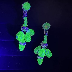 May include: A pair of dangle earrings with green glass beads. The earrings are made of metal and have a teardrop shape. The beads are iridescent and glow under a black light.