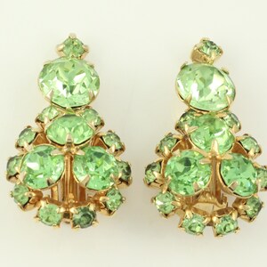 Peut inclure: Une paire de boucles d'oreilles clips avec un groupe de strass vert clair. Les boucles d'oreilles sont dor&eacute;es et ont une forme de goutte.