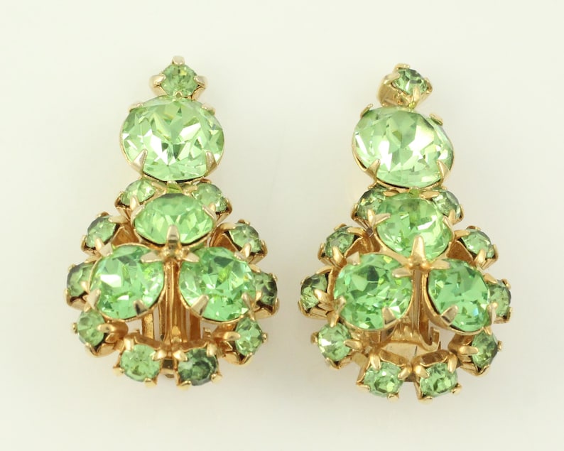 Peut inclure: Une paire de boucles d'oreilles clips avec un groupe de strass vert clair. Les boucles d'oreilles sont dor&eacute;es et ont une forme de goutte.